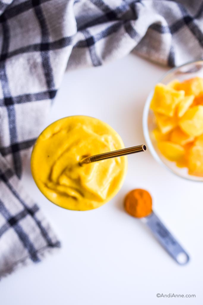 blended mango turmeric ginger smoothie, looking down at it with dark gold straw. A bowl of mango, spoon with turmeric and striped kitchen towel surround it