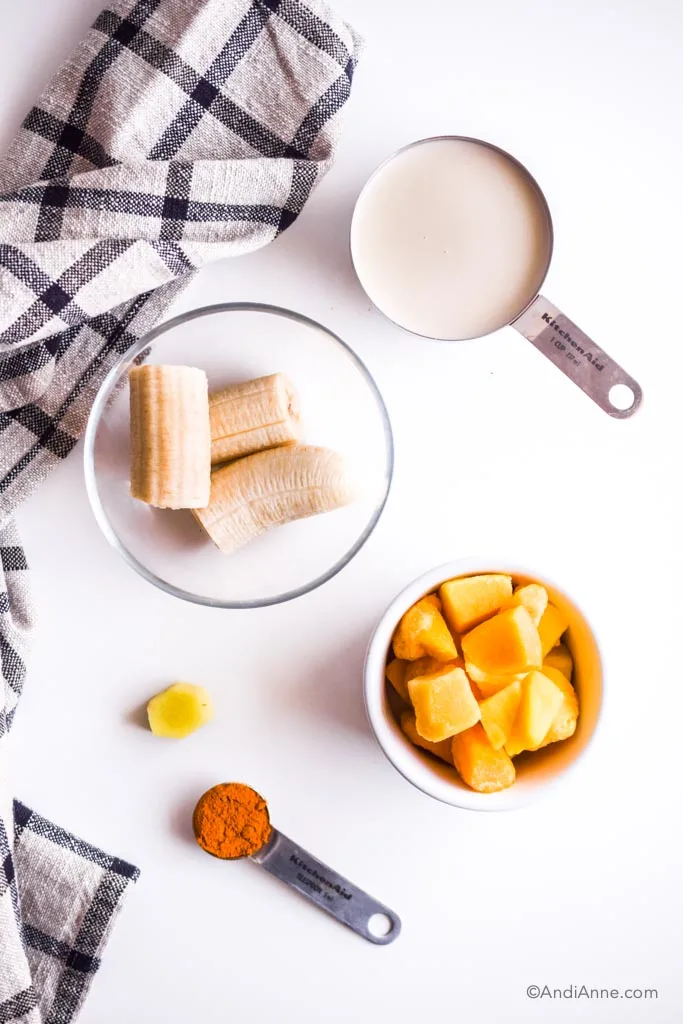 bowl of chopped frozen banana, 1 cup milk, bowl of frozen mango, piece of fresh ginger, spoon of turmeric powder on white table with striped black towel