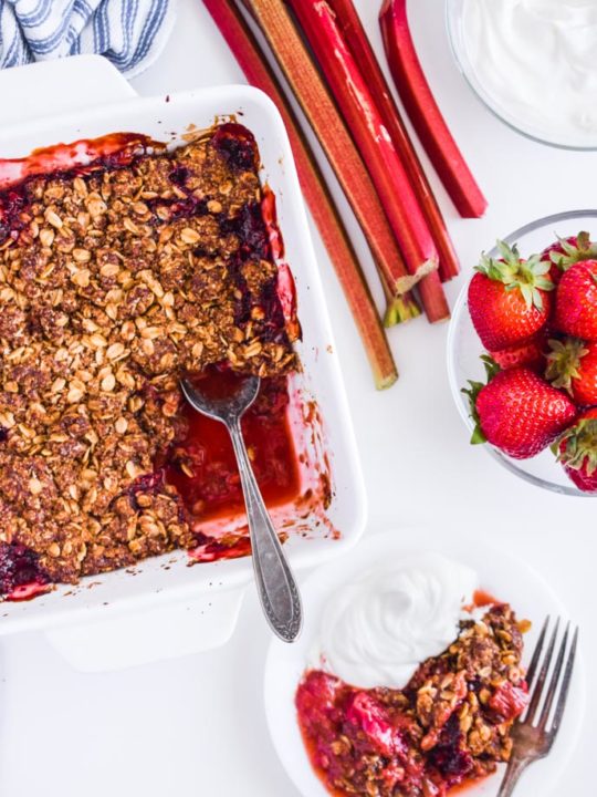strawberry rhubarb crisp in white baking dish with a piece taken out. Small plate of crisp along with a scoop of yogurt and fork. Fresh strawberries in a bowl and sticks of rhubarb are beside it.