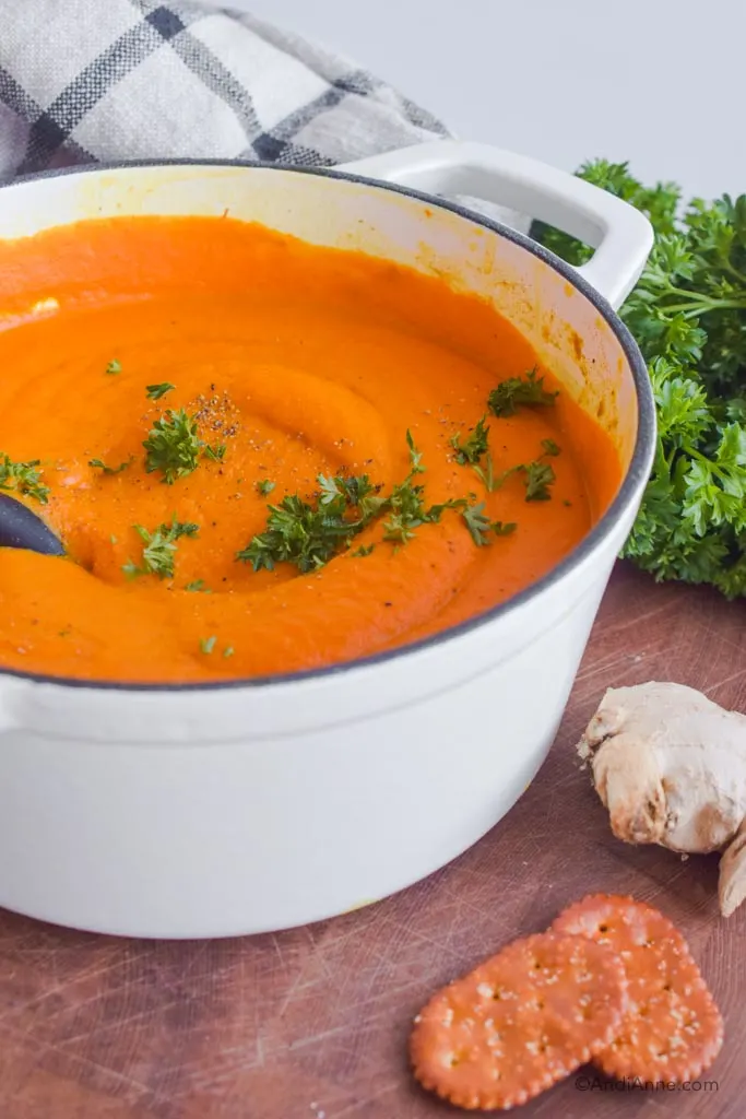 Side angle of white dutch oven with turmeric ginger carrot soup inside. Crackers, a chunk of fresh ginger and parsley are all beside the bowl.