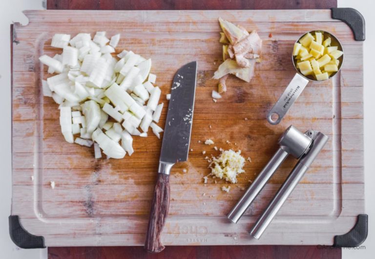 chopped onion, chopped fresh ginger in a measuring cup, a knife, a garlic mincer and minced garlic all on a cutting board.