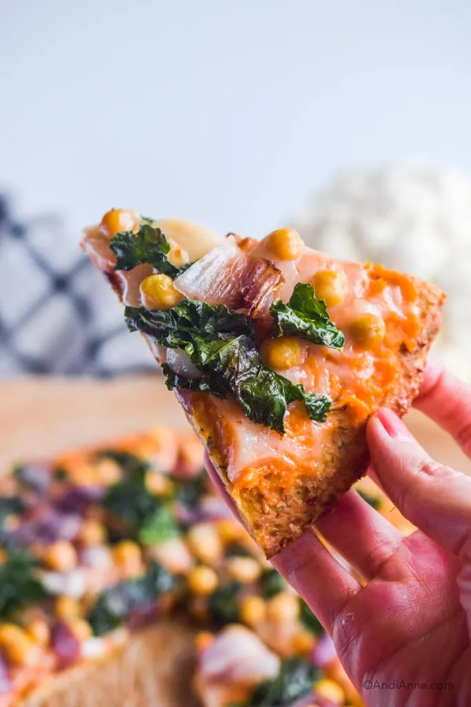 hand holding up a slice of kale chickpea cauliflower pizza with whole pizza in background