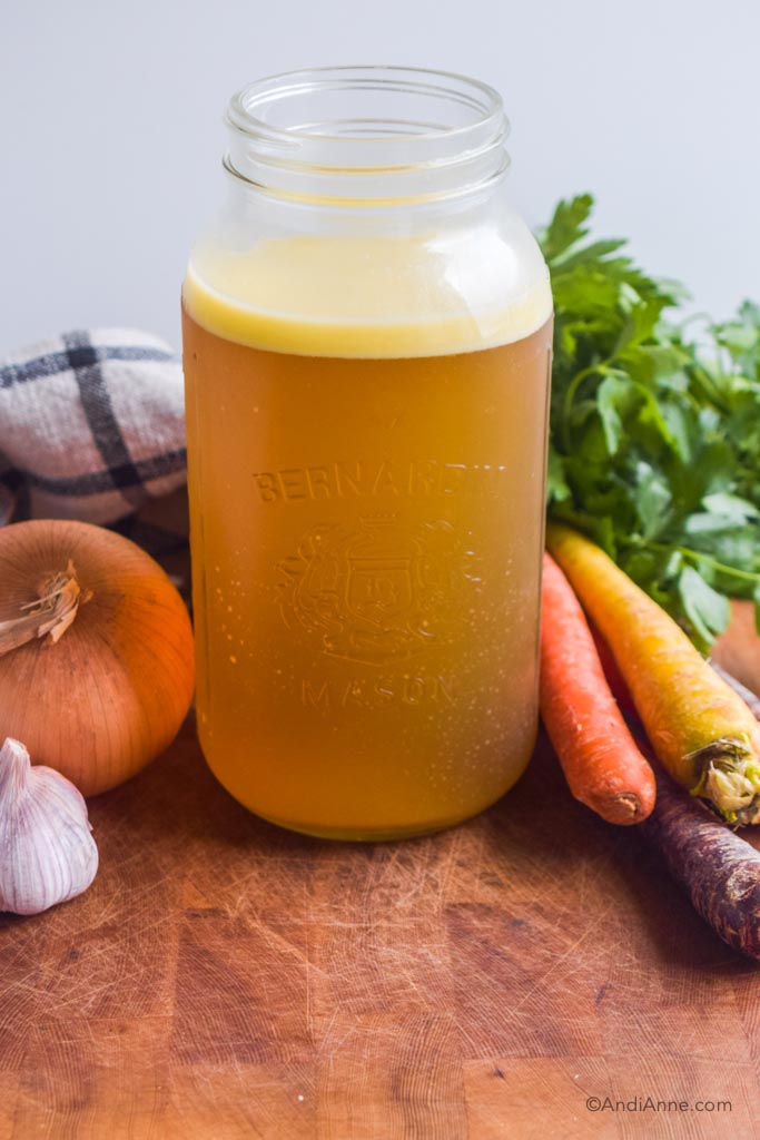 homemade bone broth in a large mason jar with carrots, parsley, onion and garlic surrounding the jar.