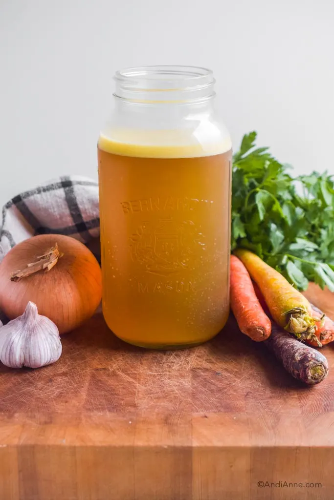 Homemade bone broth in a large mason jar with raw vegetables and a kitchen towel surrounding it.