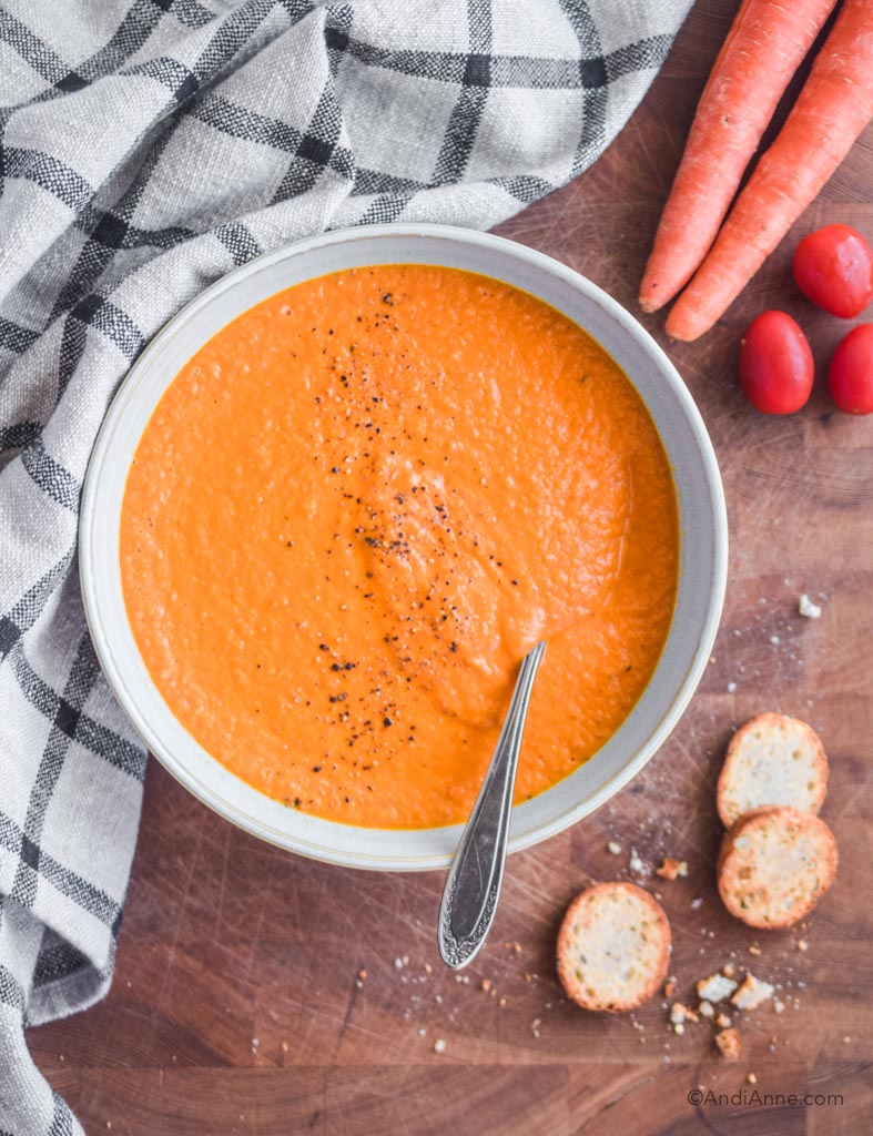 carrot tomato soup in bowl with spoon. A kitchen towel, carrots and tomatoes, and crumbled crackers surround the bowl.