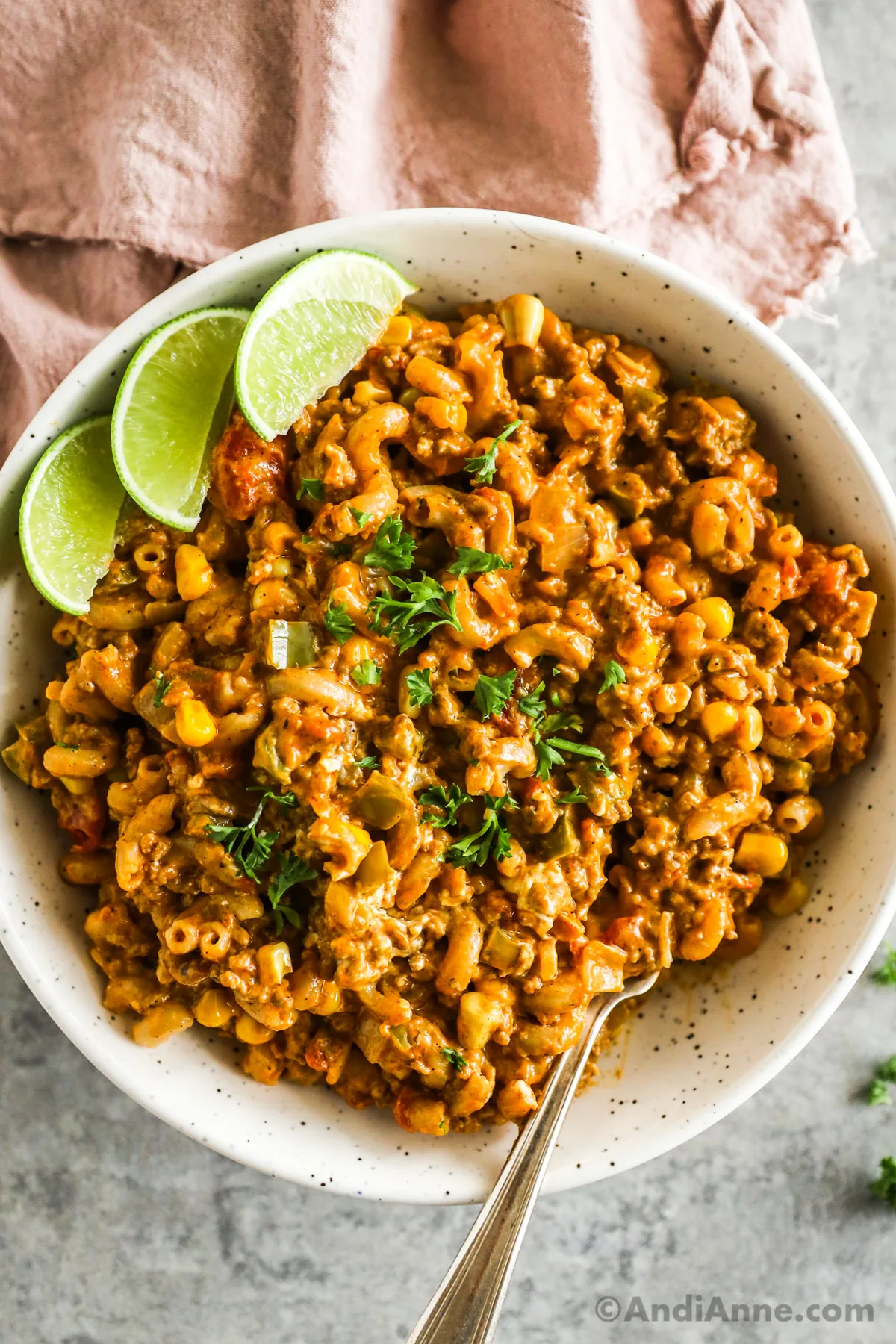 A bowl of taco pasta with lime wedges and chopped parsley  and a fork.