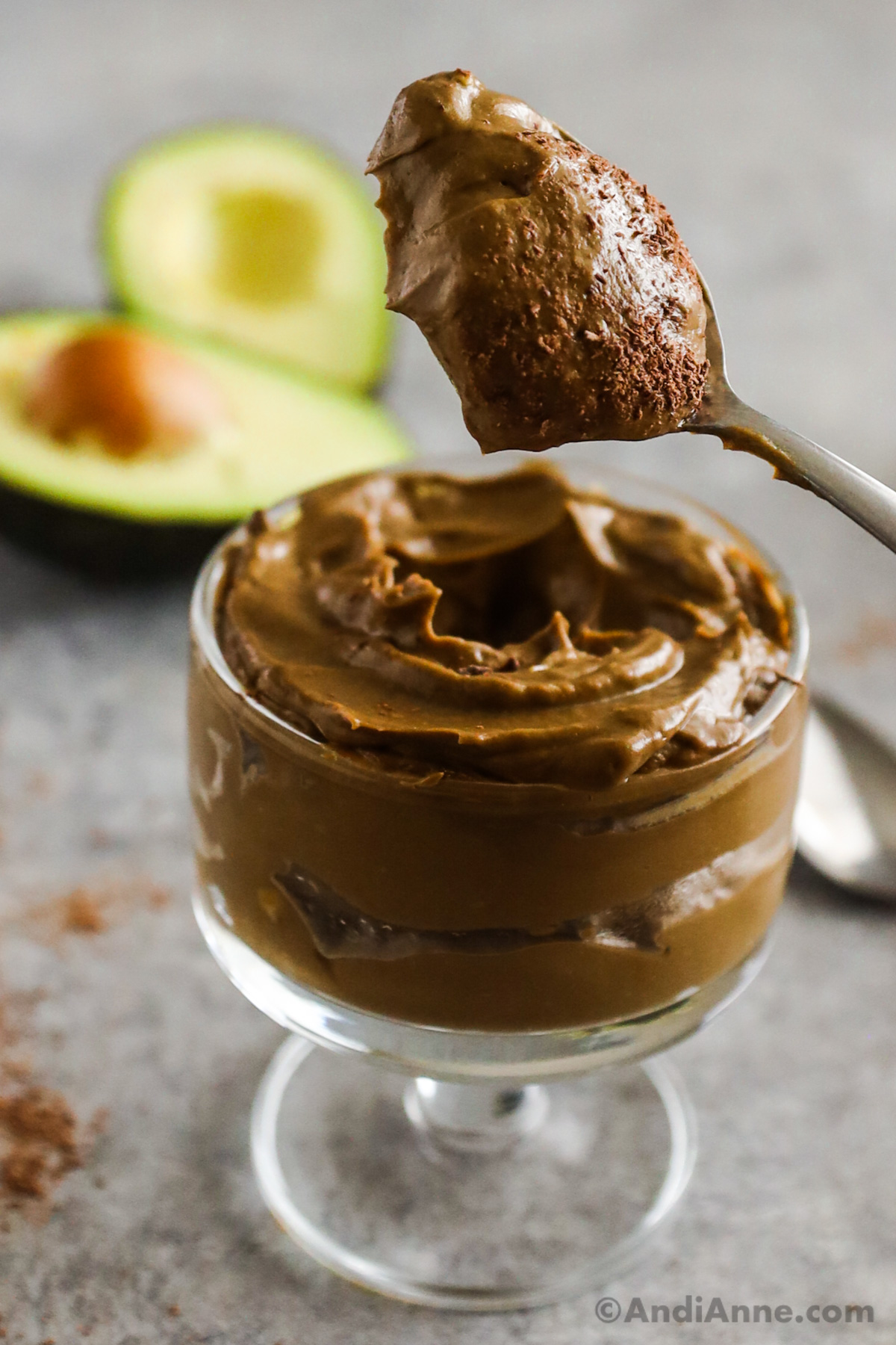A spoon with chocolate pudding above the serving bowl with sliced avocados in the background.