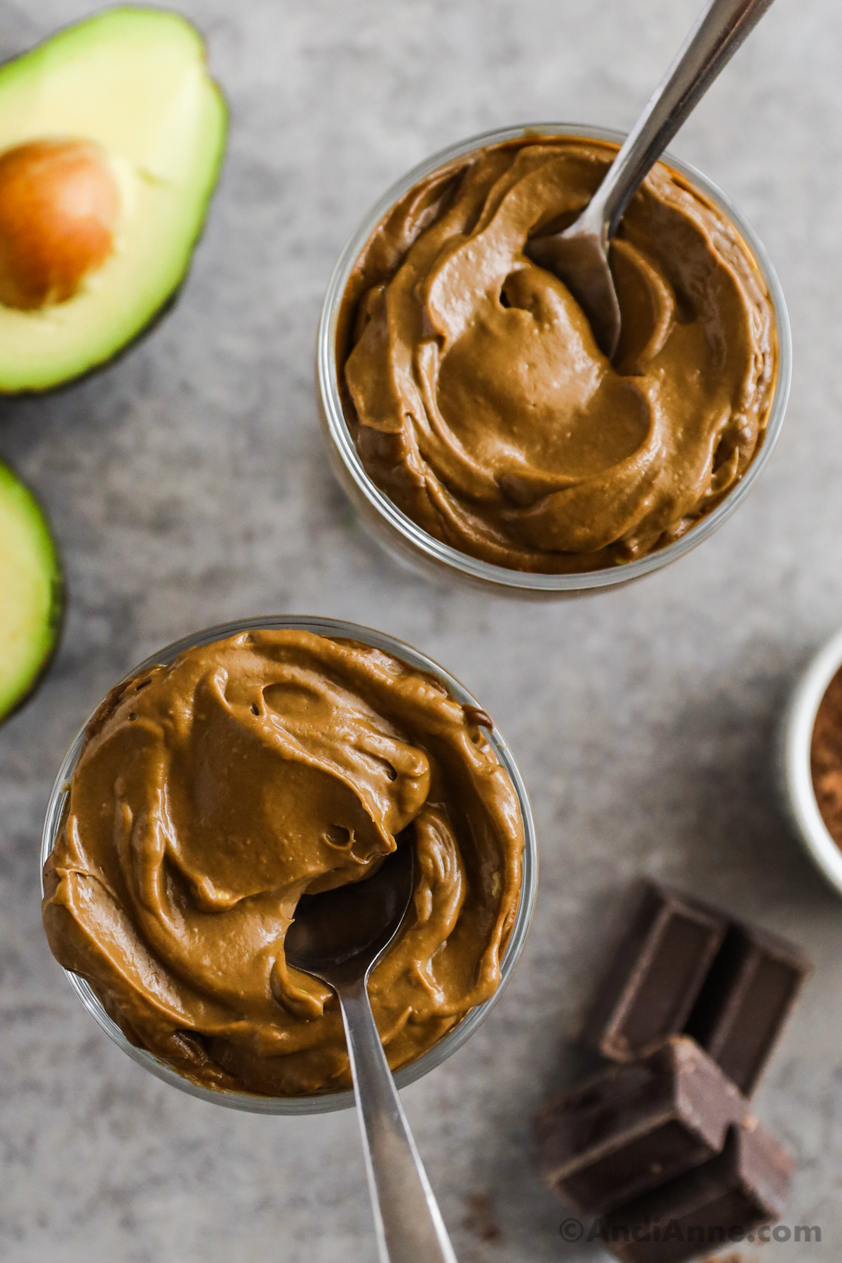 Two bowls with chocolate avocado pudding with chocolate squares and a sliced avocado beside it.