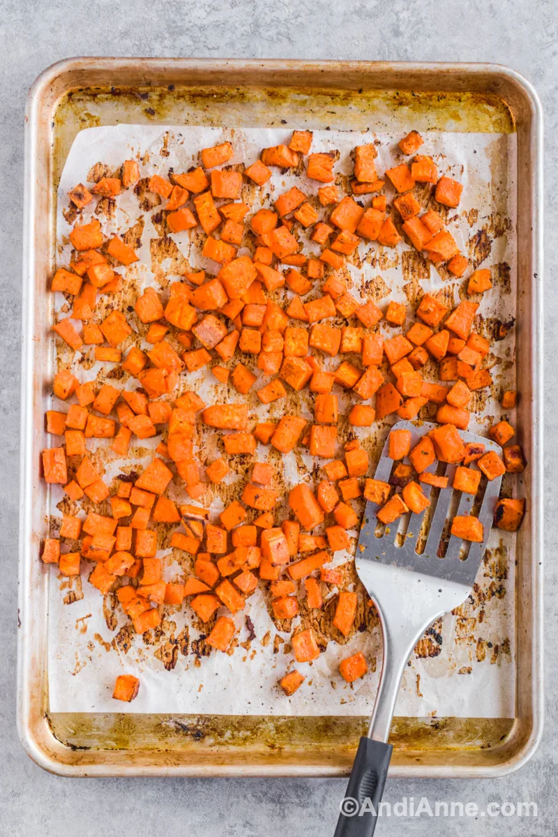 A baking sheet with baked sweet potato cubes and a metal spatula.