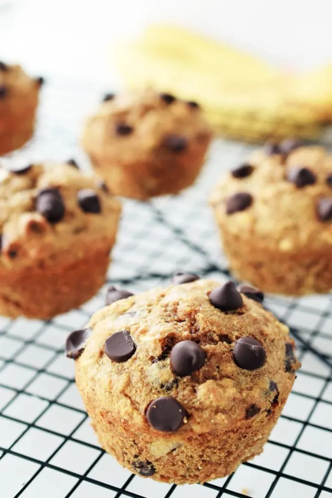 spelt muffin with banana and chocolate chips on a black cooling rack. bananas blurred in background.