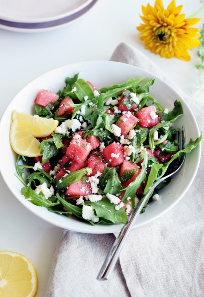 watermelon arugula salad in a white bowl surrounded by flowers