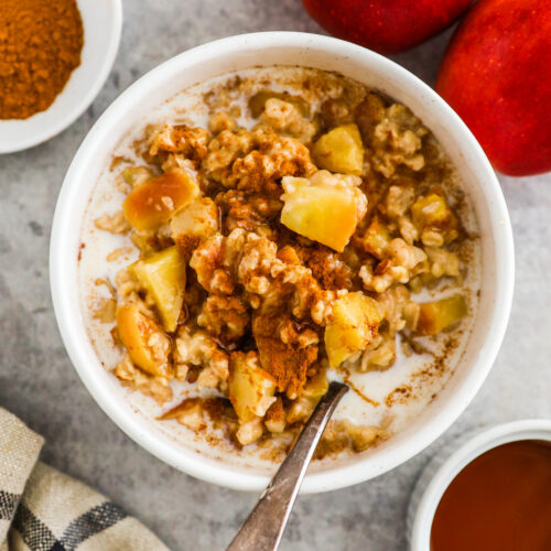 A bowl of apple cinnamon oatmeal with a spoon, surrounded by fresh red apples and spices.