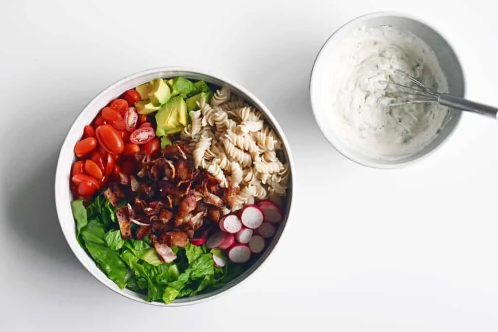 Tomatoes, bacon, pasta noodles, radish and lettuce in a large bowl, with premixed salad dressing in a smaller bowl with whisk