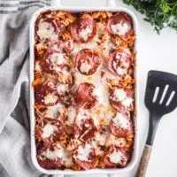 White casserole dish with pizza pasta casserole recipe, a kitchen towel and spatula beside it.