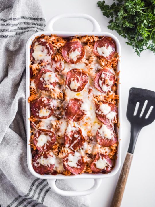 pizza pasta bake in white casserole dish with spatula, parsley and kitchen towel surrounding it