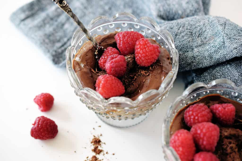 chocolate avocado pudding in two glass bowls with fresh raspberries on top and spoon. Grey napkin in background
