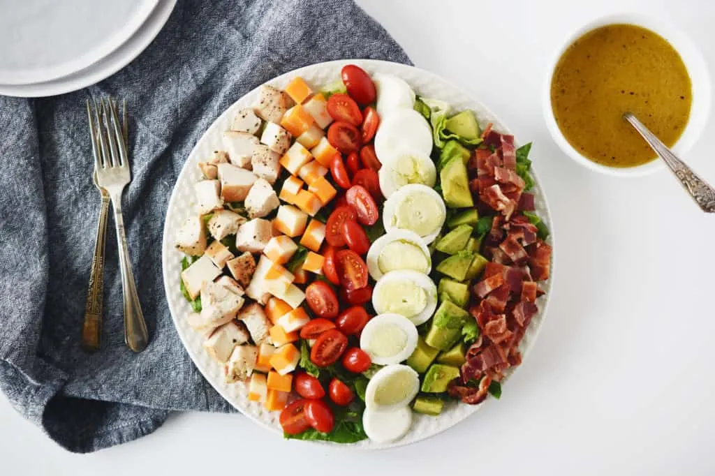 cobb salad on white plate with forks, dark napkin and white bowl of salad dressing beside the plate