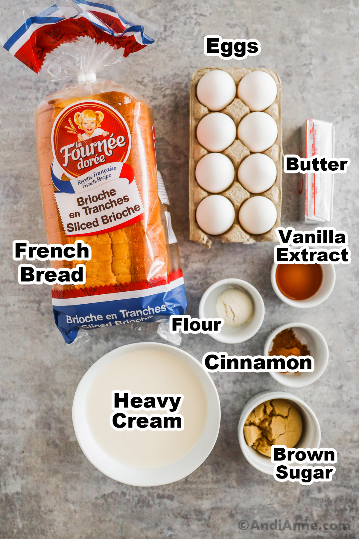Recipe ingredients on the counter including a loaf of bread, eggs in a carton, stick of butter, and bowls of vanilla extract, flour, cinnamon, heavy cream and brown sugar.