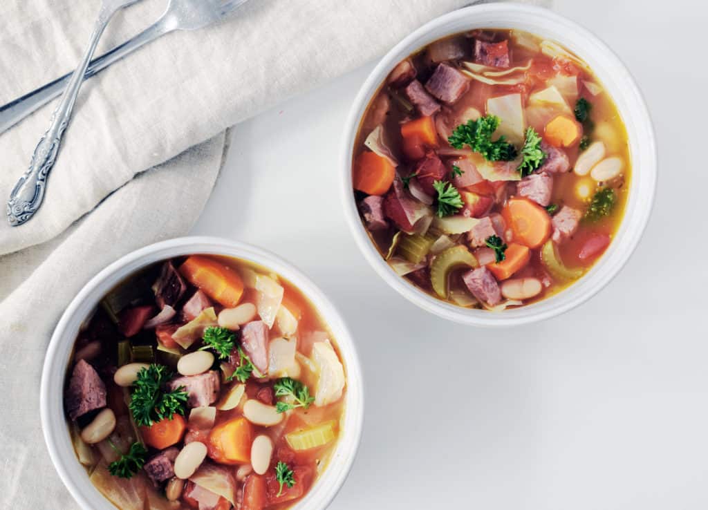 ham bean and cabbage soup in two white serving bowls with spoons