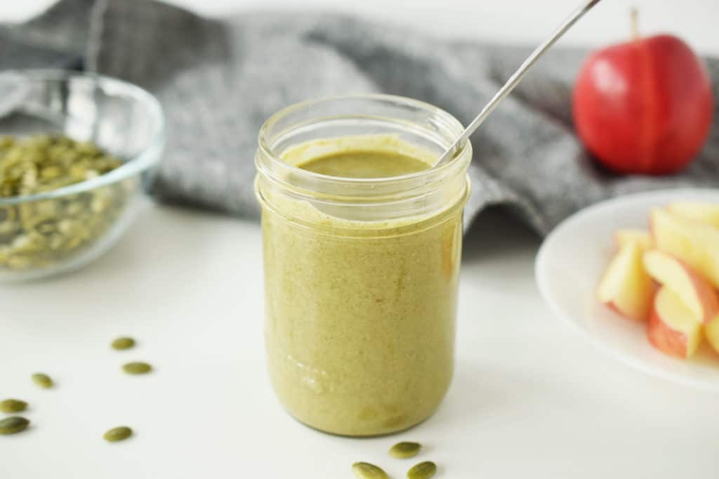 homemade pumpkin seed butter in a mason jar with spoon, surrounded by apple slices and pumpkin seeds. 