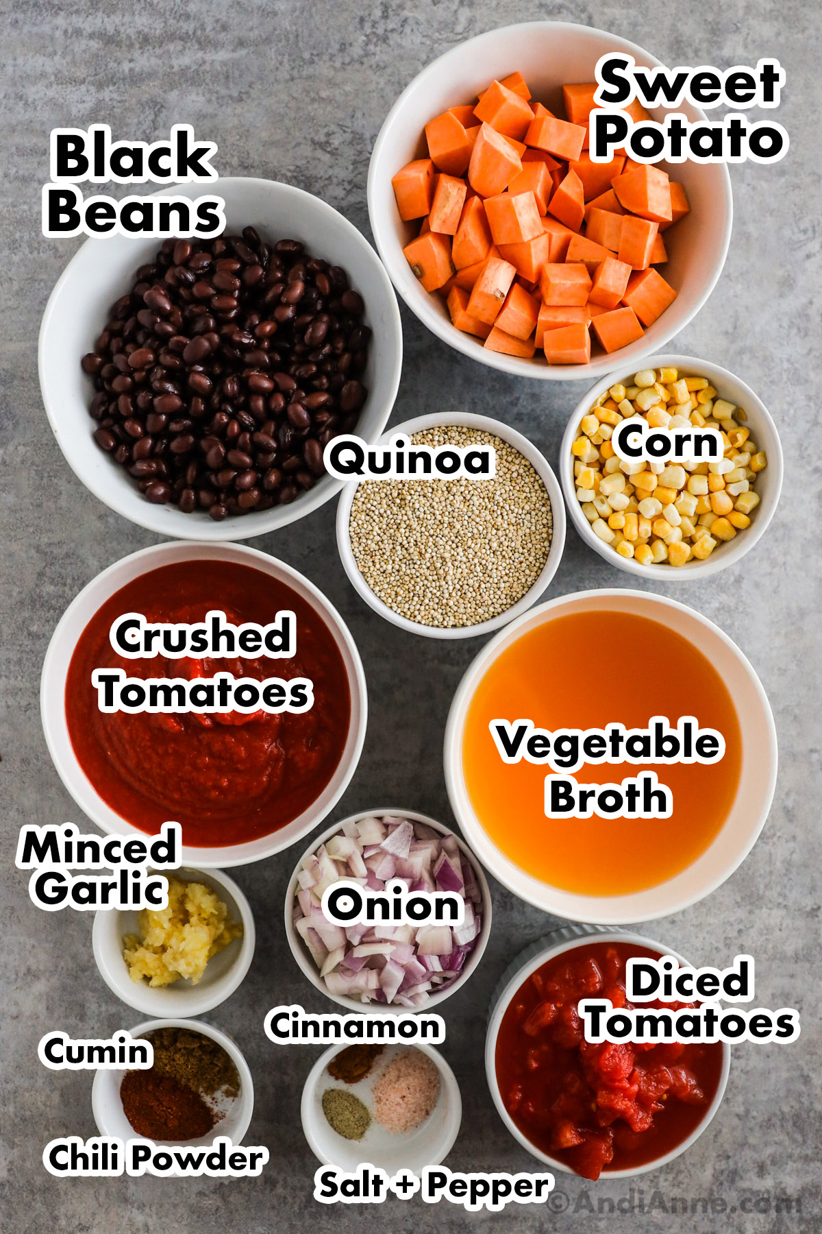 Recipe ingredients on the counter including bowls of chopped sweet potato, black beans, raw quinoa, corn, crushed tomatoes, vegetable broth, minced garlic, chopped onion, diced tomatoes, and spices.