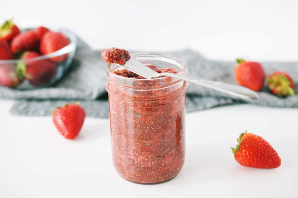 strawberry chia jam in mason jar with spoon. fresh strawberries in a bowl and dark grey napkin are in background.