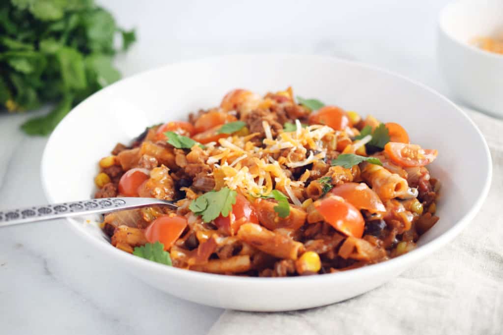 taco pasta in white bowl with silver spoon. Green parsley in background