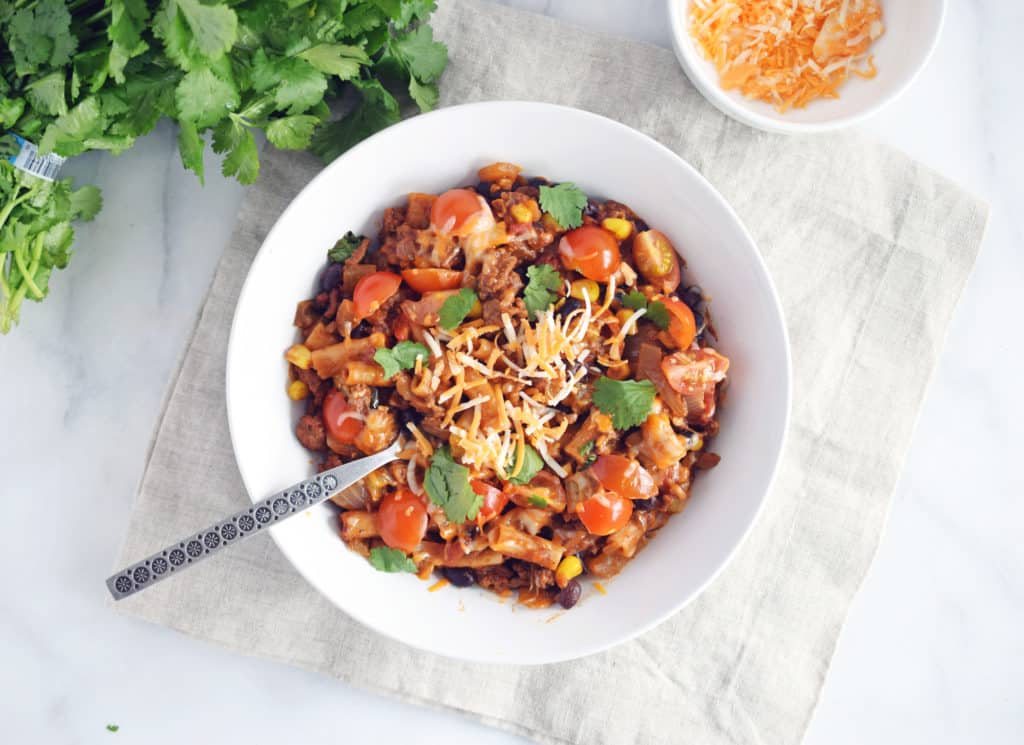 one pot taco pasta in a white bowl with silver spoon on beige napkin. Parsley and shredded cheese in the top of picture.