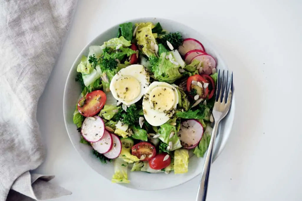 garden salad with two hard boiled eggs on a white plate with fork and beige napkin on left