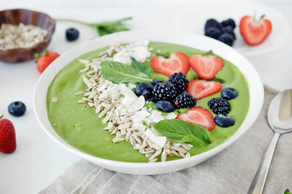 close up of tropical green smoothie in white bowl with silver spoon and berries and background.