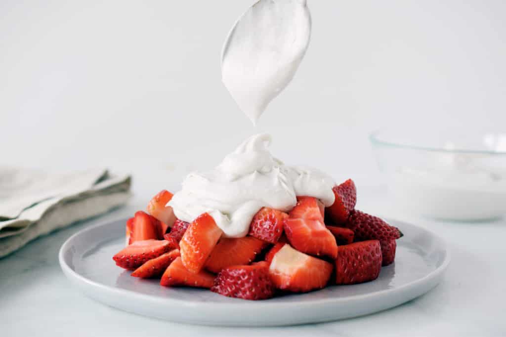 whipped coconut cream being spooned on top of sliced strawberries on a grey plate