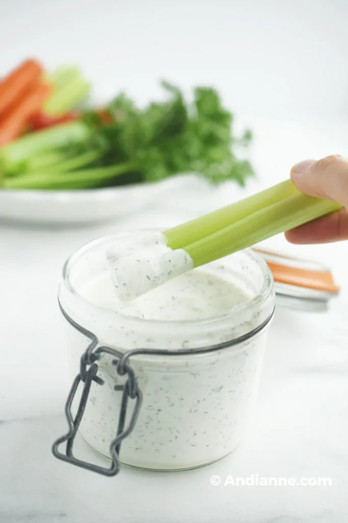 dipping celery into homemade buttermilk ranch dressing in a jar with vegetables in background