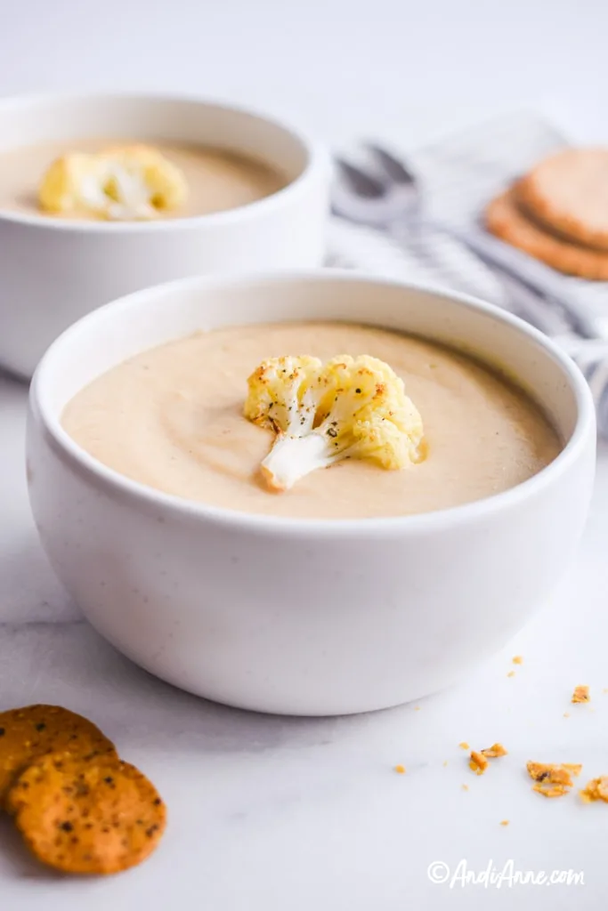 close up of white bowl with cauliflower soup recipe inside it. One piece of roasted cauliflower on top and crackers crumbled below bowl.