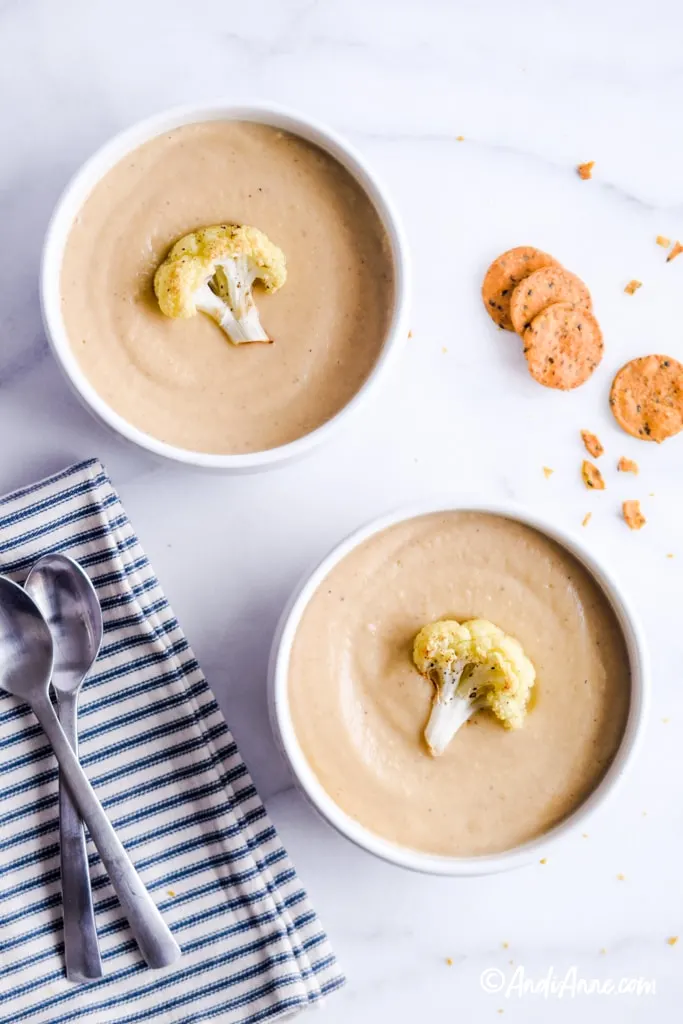 cauliflower soup in white bowls with one piece of roasted cauliflower on top. Two spoons on a striped napkin beside it.