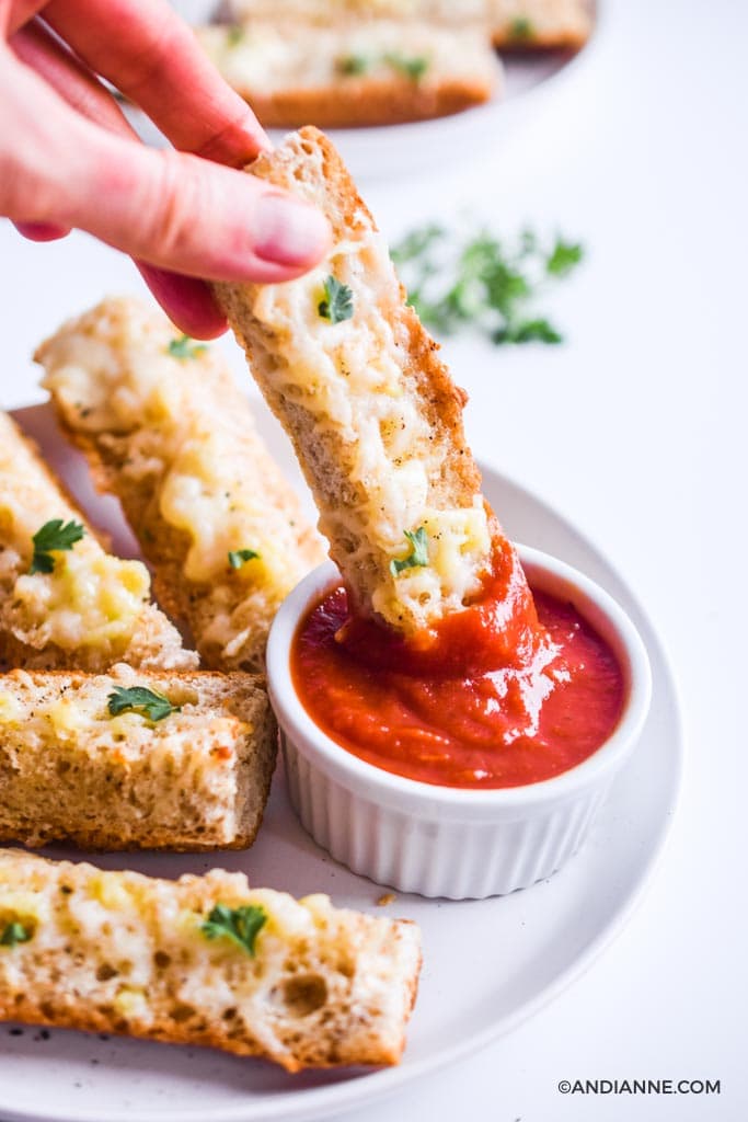 fingers dipping a cheesy garlic french bread stick into small white cup of pizza sauce