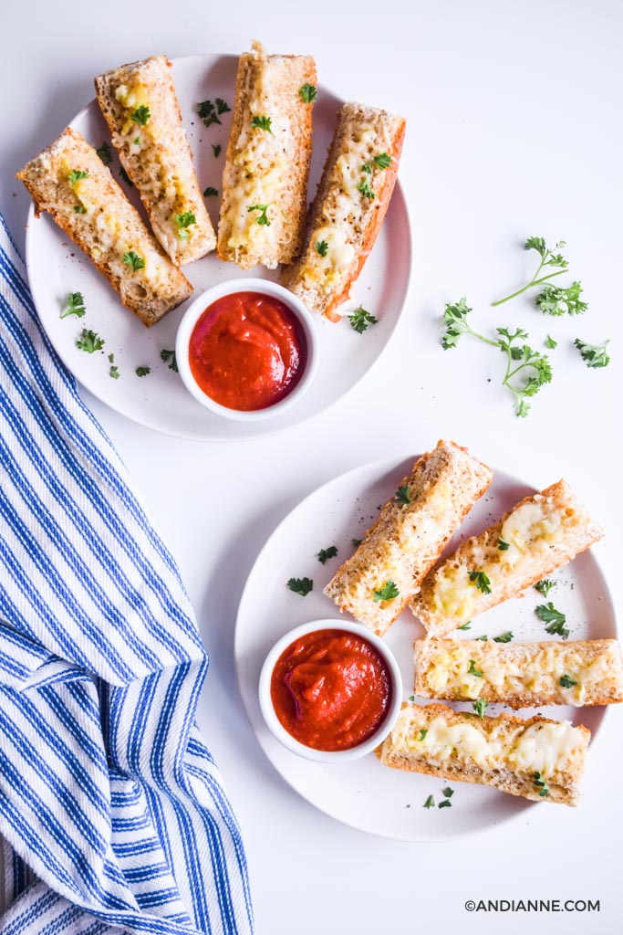 slices of cheesy garlic french bread sticks on white plates with small cups of pizza sauce. Blue striped napkin and bits of parsley on counter.
