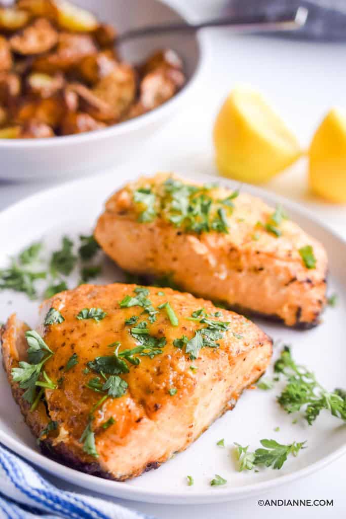 air fryer salmon on white plate sprinkled with chopped parsley. lemons and a bowl of potatoes in background