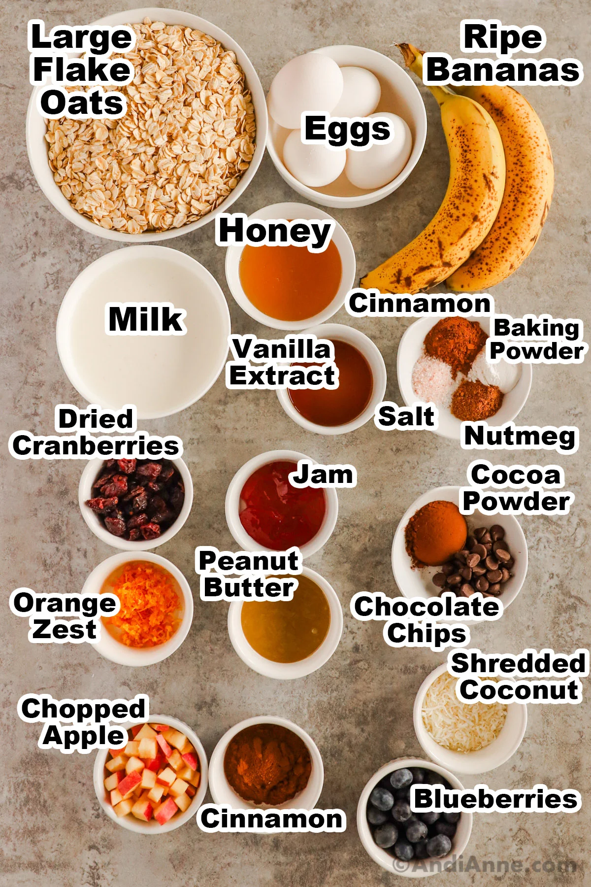 Recipe ingredients in bowls on counter including bowl of large flake oats, eggs, bananas, honey, milk, cinnamon, spices, jam, dried cranberries, peanut butter, chocolate chips, shredded coconut, apple, cinnamon and shredded coconut.