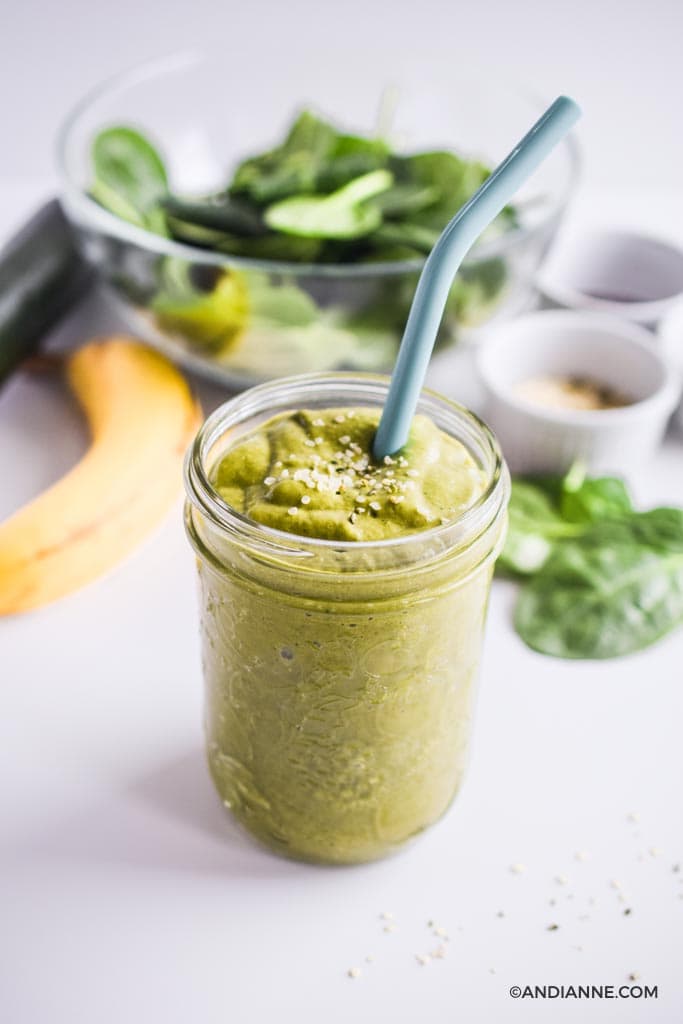 healthy green smoothie in a mason jar with blue straw surrounded by banana, spinach leaves and small white bowls