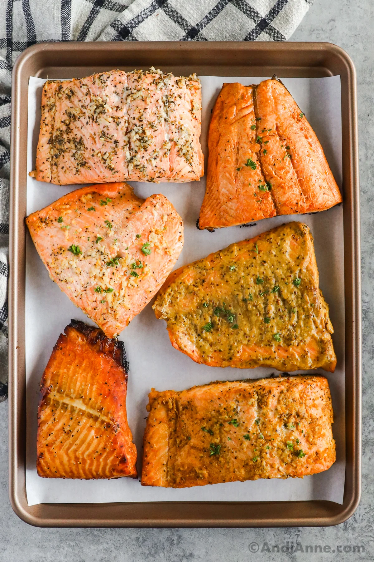 Six cooked salmon fillets coated in different seasonings on a baking sheet.