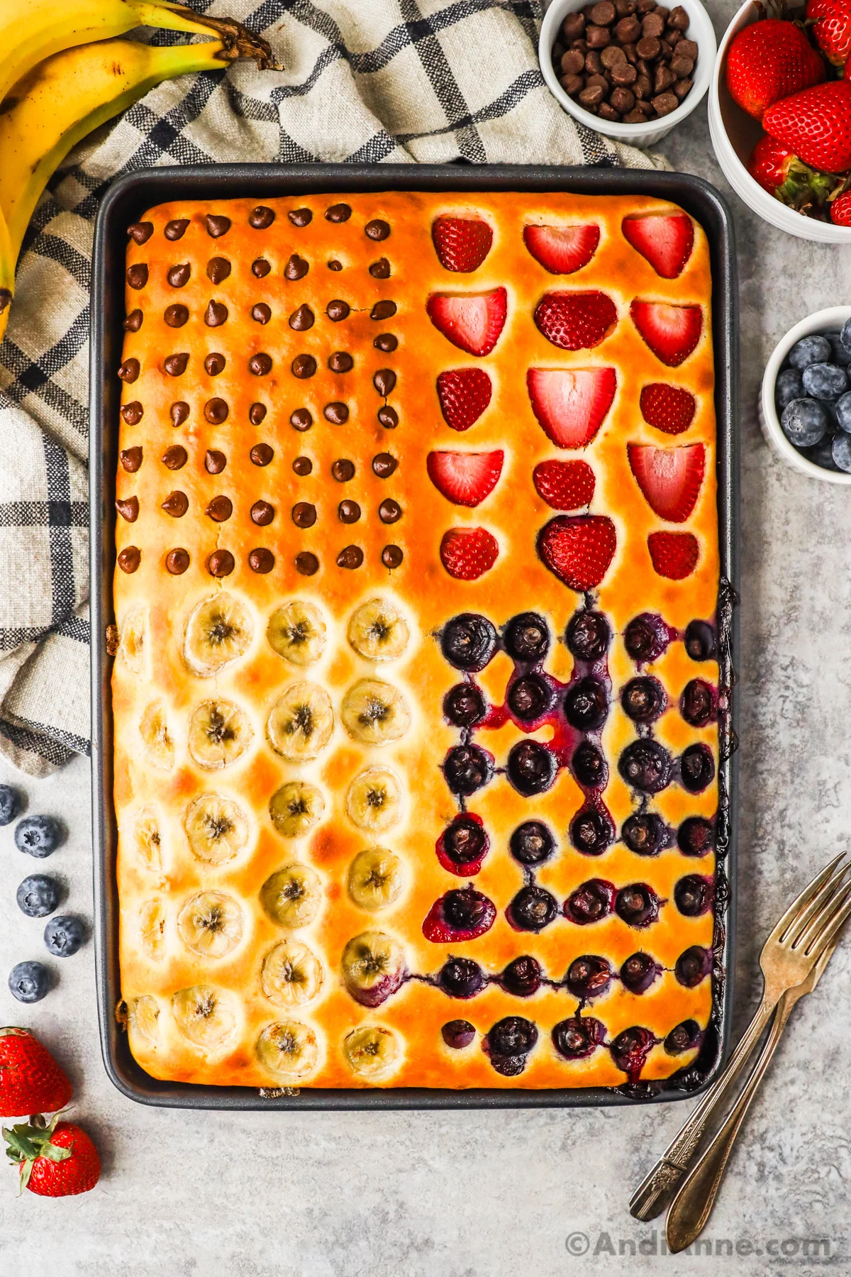 A baking sheet with cooked sheet pan pancake inside topped with blueberries, sliced bananas, strawberries, and chocolate chips.