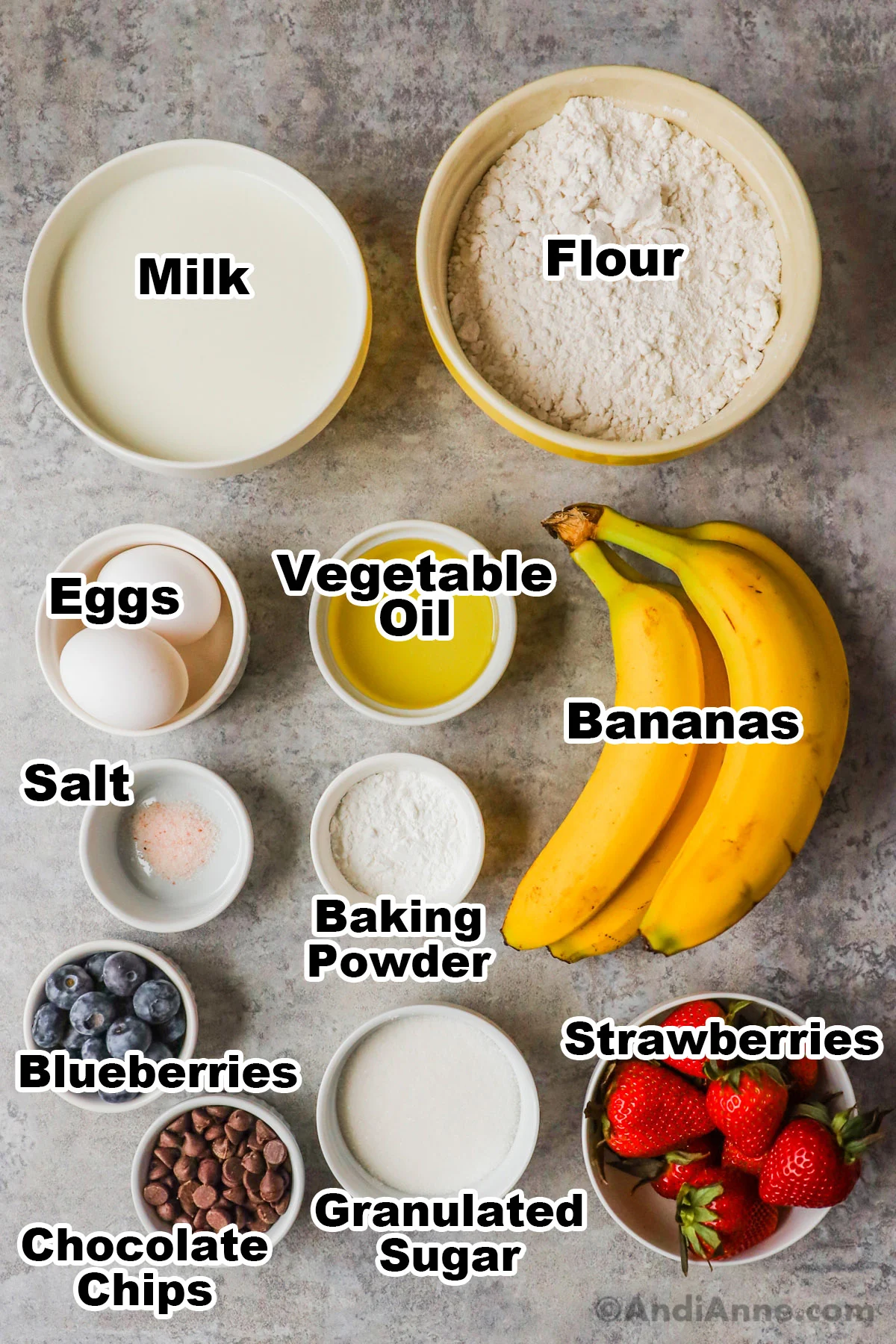 Recipe ingredients including bowls of milk, flour, eggs, vegetable oil, salt, baking powder, blueberries, bananas, strawberries and sugar.