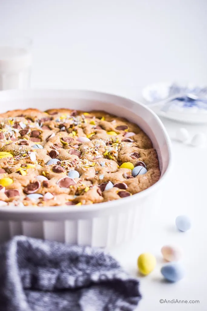 side angle of white casserole dish with cooked blondies inside. Plate and glass of milk are blurred in background.