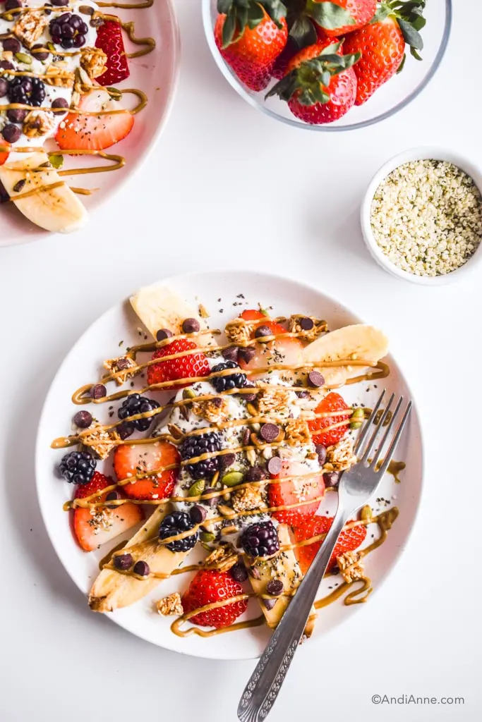 healthy banana splits on pink plates with a fork. small bowls of hemp seeds and fresh strawberries beside them.