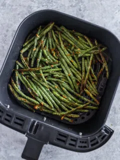 An air fryer basket with green beans and crispy small pieces of parmesan cheese.
