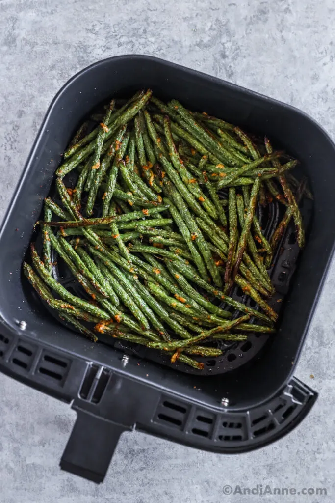 An air fryer basket with green beans and crispy small pieces of parmesan cheese.