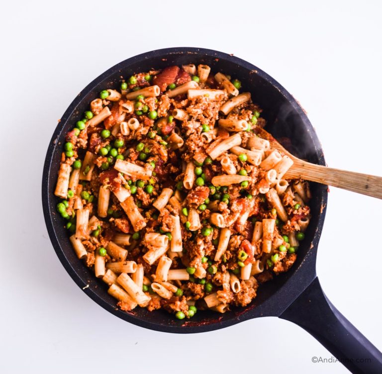 cooked pasta with peas and ground chicken in a black frying pan with wood spoon