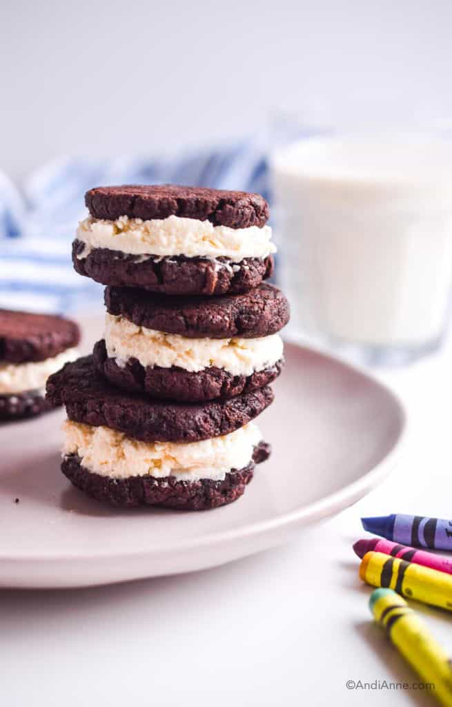 homemade oreos stacked in three on a pink plate. Four colored crayons are in the foreground. A glass of milk in the background.