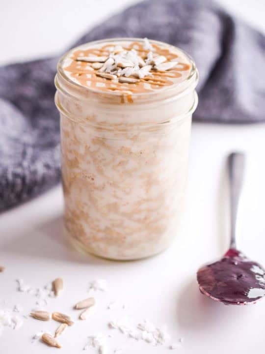 oats in mason jar with spoon of red jam beside it. seeds and nuts on sprinkled on to counter.