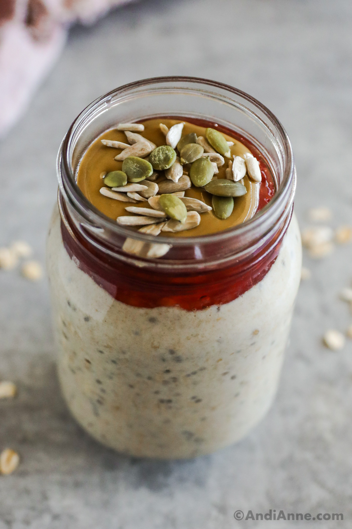 A jar of peanut butter overnight oats with jam, peanut butter and seeds on top.